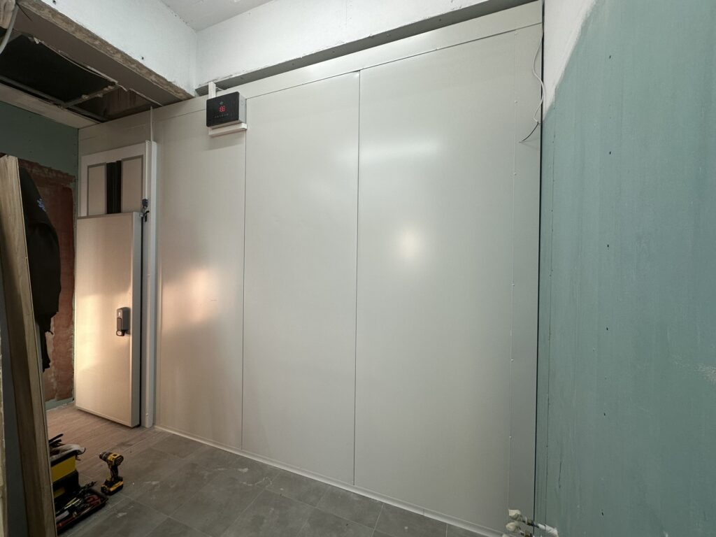 Βιομηχανικός ψυκτικός θάλαμος με πόρτα ασφαλείας και σύστημα ελέγχου θερμοκρασίας, εγκατεστημένος σε επαγγελματικό χώρο από εξειδικευμένη ψυκτική εταιρεία.